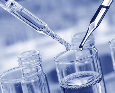 Close-up of two pipettes above a glass container filled with liquid, demonstrating a laboratory setting.