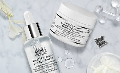 Alt text: A flat lay of skincare products from Kiehl's, including a dropper bottle of Clearly Corrective Dark Spot Solution and a jar of Clearly Corrective Brightening & Smoothing Moisture Treatment, arranged on a marble surface with white flowers and leaves.