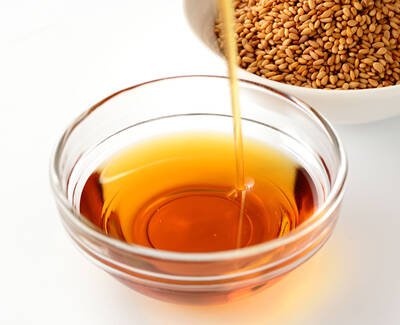 A glass bowl filled with golden-brown sesame oil, with a stream of oil pouring into it from above. In the background, a white bowl contains dry sesame seeds.