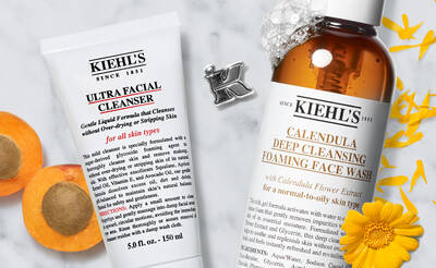 Alt text: Two Kiehl's facial cleansers are displayed on a marble surface, accompanied by apricots and yellow flower petals. The left product is a tube labeled "Ultra Facial Cleanser" and the right is a bottle labeled "Calendula Deep Cleansing Foaming Face Wash."