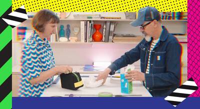 A woman and a man engage in a hands-on activity at a table, with various craft supplies around them. The woman holds a small black bag, while the man pours liquid from a carton into a bowl. The background features colorful patterns and designs.
