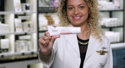 A smiling woman with curly hair holds a tube of Kiehl's cream in a retail setting, surrounded by skincare products on shelves.