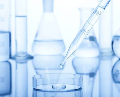 A clear pipette is poised above a petri dish with a droplet about to fall, surrounded by laboratory glassware in shades of blue.