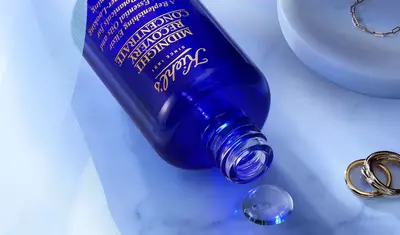 A blue bottle of Tatcha's Midnight Concentrate skincare product, partially spilled on a marble surface, with a crystal-like droplet nearby and some gold rings in the background.