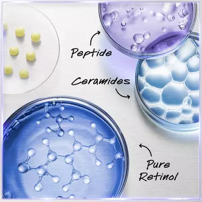 Alt text: A close-up of three petri dishes containing different skincare ingredients: one with a purple liquid labeled "Peptide," another with a blue gel labeled "Pure Retinol," and the third with a clear substance labeled "Ceramides." Small yellow spheres are arranged on a white surface beside the dishes.