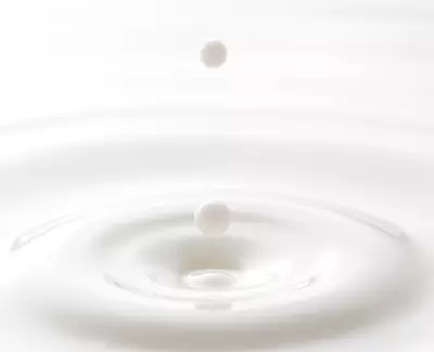 A drop of liquid is about to fall into a smooth, white surface, creating ripples in the water below.