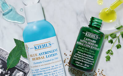 Two Kiehl's skincare products are displayed: a blue bottle of Blue Astringent Herbal Lotion and a green bottle of Cannabis Sativa Seed Oil Herbal Concentrate, surrounded by herbal ingredients and drops of oil.