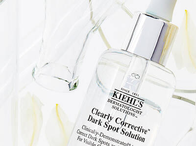 Bočica Kiehl's Clearly Corrective™ Dark Spot Solutiona među bijelim cvjetovima i staklenim predmetima.