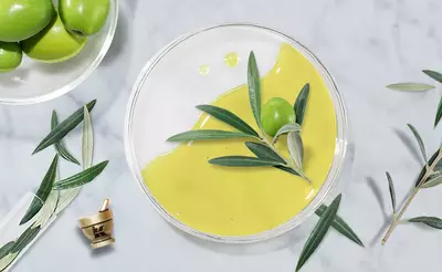 A clear bowl filled with a smooth, green mixture sits on a marble surface, accompanied by fresh green olives and olive leaves. A small mortar and pestle is placed nearby, suggesting an olive oil preparation theme.