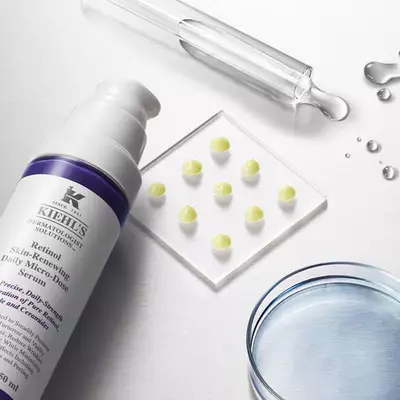 A skincare product with a pump dispenser, accompanied by a glass pipette, a petri dish containing small yellow serum droplets, and droplets of water on a smooth surface.