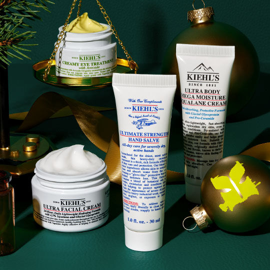 A festive arrangement of Kiehl's products including Creamy Eye Treatment on golden scales, Ultra Facial Cream, Ultimate Strength Hand Salve, and Ultra Body Mega Moisture Squalane Cream, surrounded by green ornaments, fir branches, and gold ribbons on a dark green background.