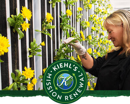 Žena s dugom kosom sadi cvijeće na verticalnoj vrtnoj stjenci, uz logo Kiehl's i natpis "Mission Renew".