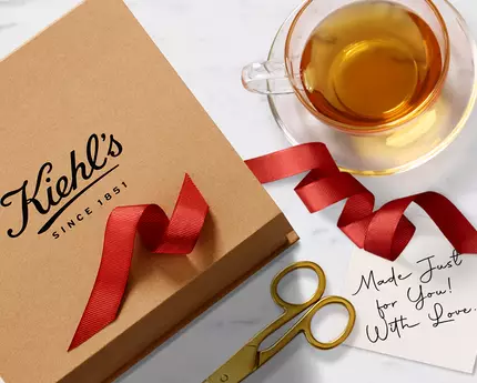 Boks s logotipom Kiehl's, crvena vrpca, škarice, šalica čaja i poruka na papiru na mramornoj površini.